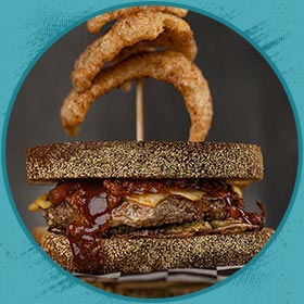 A Florida burger with dark bread, BBQ sauce, cheese, and onion rings on a skewer behind.