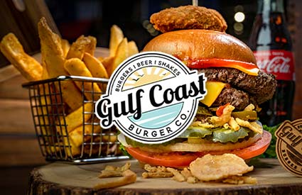 Burger with fries, a shake, and a drink behind a Gulf Coast Burger Co. logo on a wooden table.
