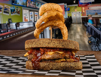 Cornbread burger with barbecue sauce, shakes, and onion rings on a checkered mat in a diner.